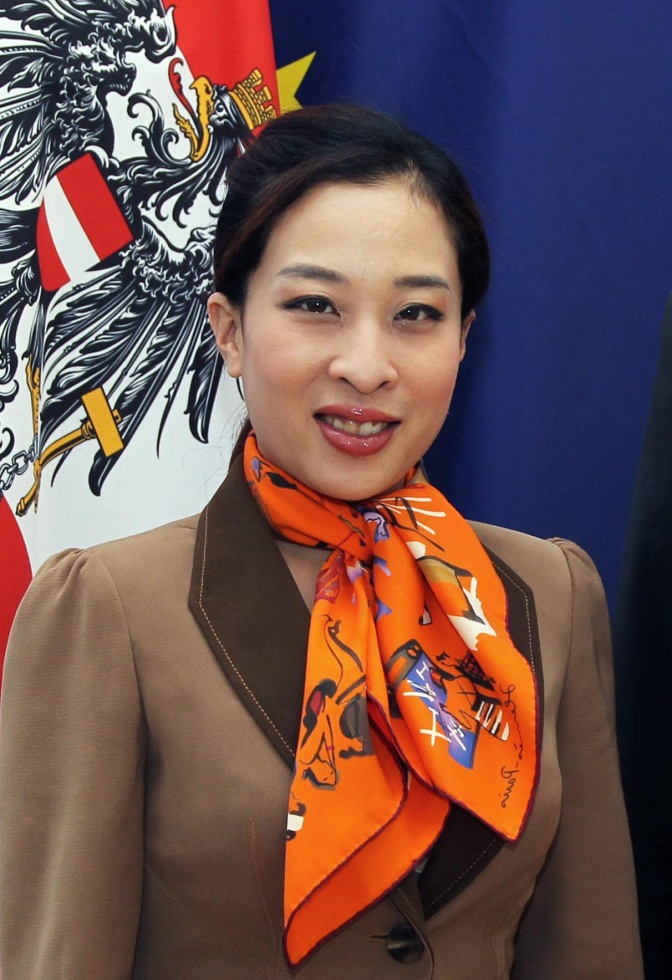 Eine junge, asiatisch aussehende Frau vor einem Blumenstrauß und einer blauen Wand. Sie trägt ein braunes Sakko und ein leuchtend orangefarbenes Halstuch.