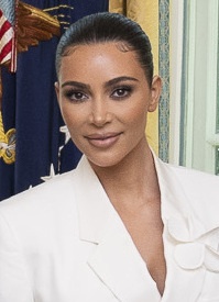Kim Kardashian mit zurückgesteckten Haaren in einem weißen Hosenanzug vor der amerikanischen Flagge.