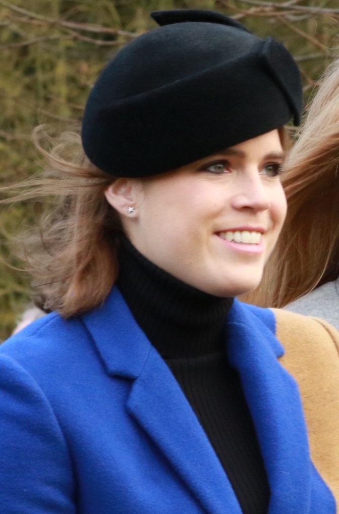 Prinzessin Eugenie in einem blauen Mantel mit schwarzem Hut. Sie hat lange braune Haare und lächelt.