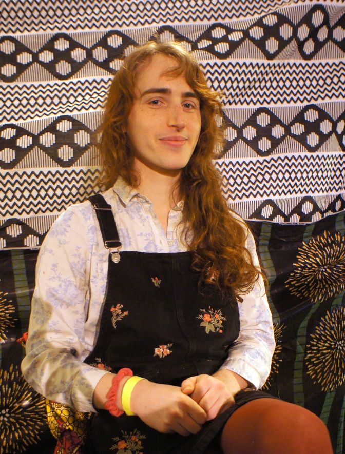 Eine trans Person in einem kurzen Cordkleid vor einer stark gemusterten Tapete