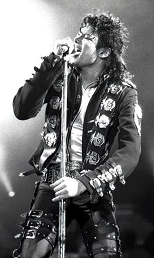 Ein schwarz-weiß-Foto von Michael Jackson in einem uniform-ähnlichen Bühnenkostüm. Er singt in ein Standmikro.