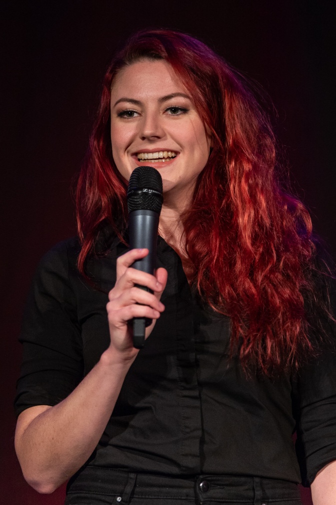 Eine schwarz gekleidete Frau mit roten Haaren spricht lächelnd in ein Mikro.