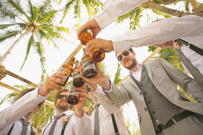 Eine Gruppe von Männern in Anzug und Sonnenbrillen stößt mit Bierflaschen miteinander an.