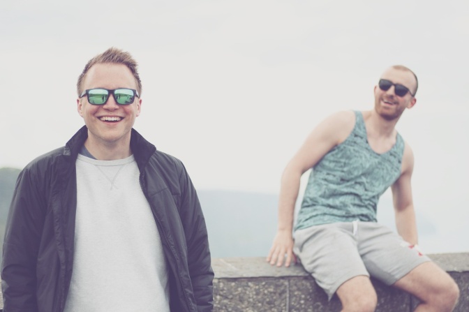 2 lachende Männer mit Sonnenbrillen vor einer Mauer