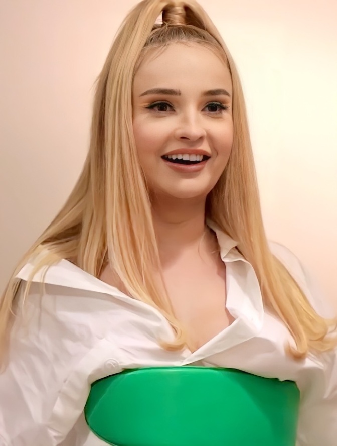 Kim Petras trägt ein grünes Bustier über einer weißer Bluse. Sie hat einen Zopf mitten auf dem Kopf gebunden und hat lange, glatte, blonde Haare.