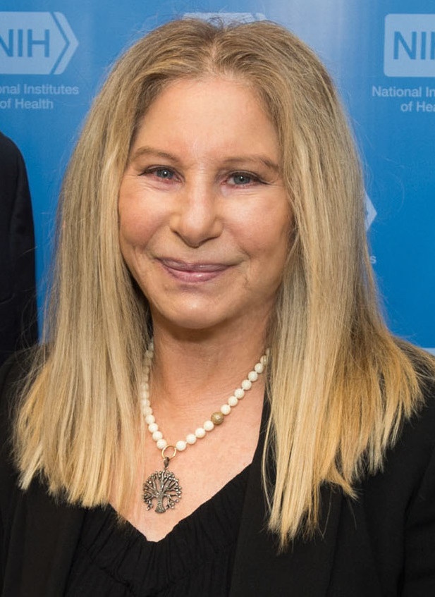 Barbra Streisand mit schulterlangen, glatten blonden Haaren. Sie trägt einen schlichten schwarzen Pulli und eine Halskette mit Anhänger.