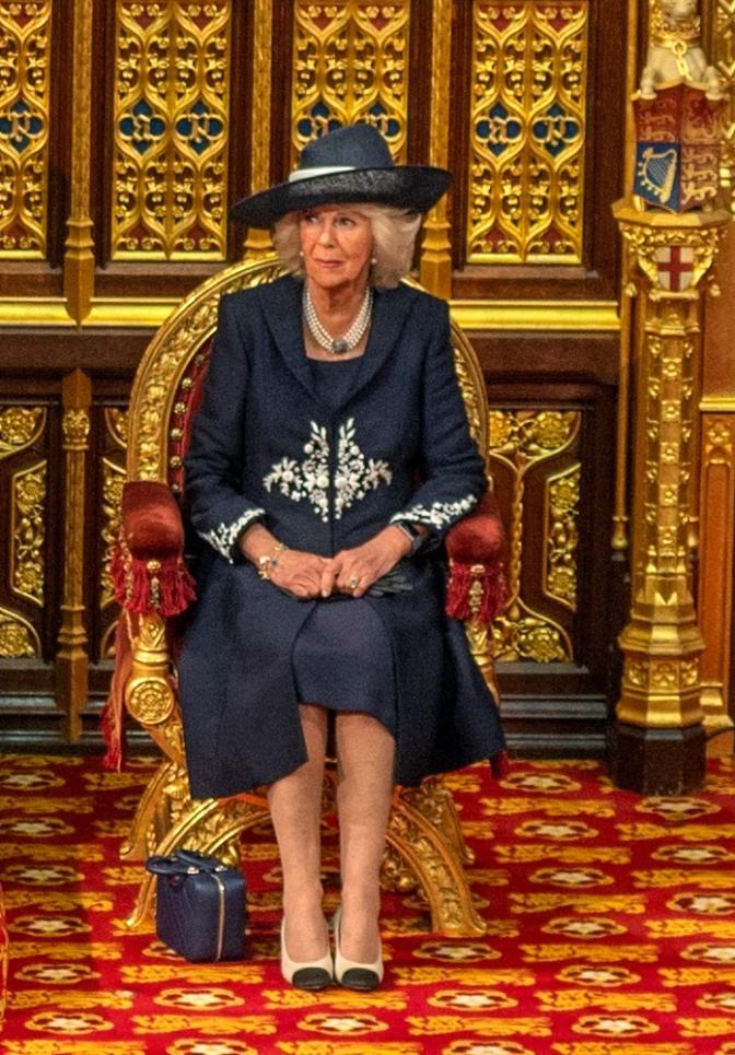 Camilla in dunkelblauem Kleid mit passendem Hut und passender Handtasche. Sie sitzt auf einem goldenen Thron vor goldener Wanddekoration.