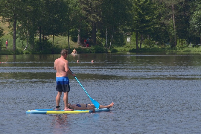 Ein Mann beim Stand up Paddling auf einem See. Vorne auf seinem Brett liegt eine Frau im Bikini.