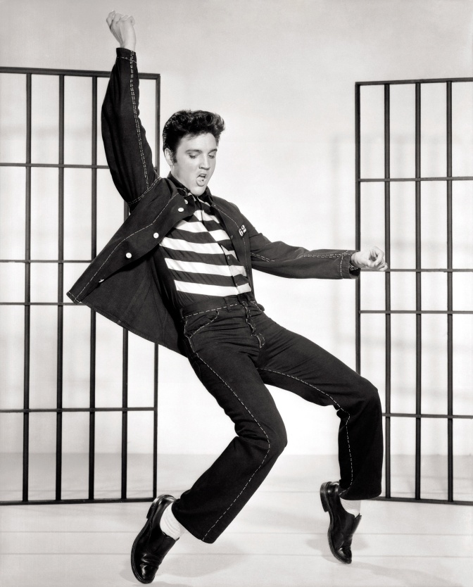 Schwarz-weiß-Foto von Elvis Presley beim Tanzen. Er steht auf Zehenspitzen und biegt den Oberkörper weit zurück. Er trägt ein T-Shirt mit Blockstreifen zu Jeans und Jeanshemd.
