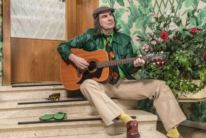 Guildo Horn spielt auf einer Akustikgitarre und sitzt auf einer Treppenstufe.