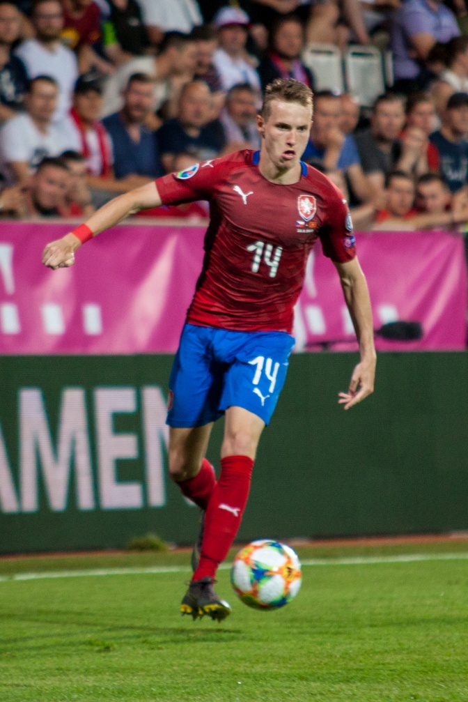 Jakub Jankto in einem roten Trikot und blauer Hose auf dem Fußballfeld. Er hat kurze blonde Haare. Er spielt gerade den Ball.