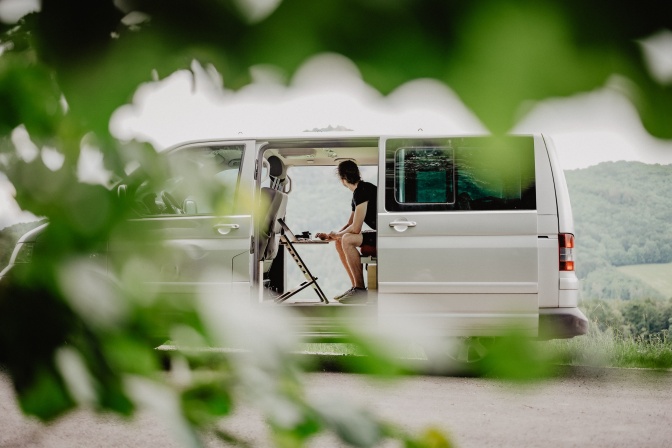 Ein weißer Van mit auf beiden Seiten geöffneten Türen steht im Freien. Darin sitzt ein Mann und arbeitet am Laptop.
