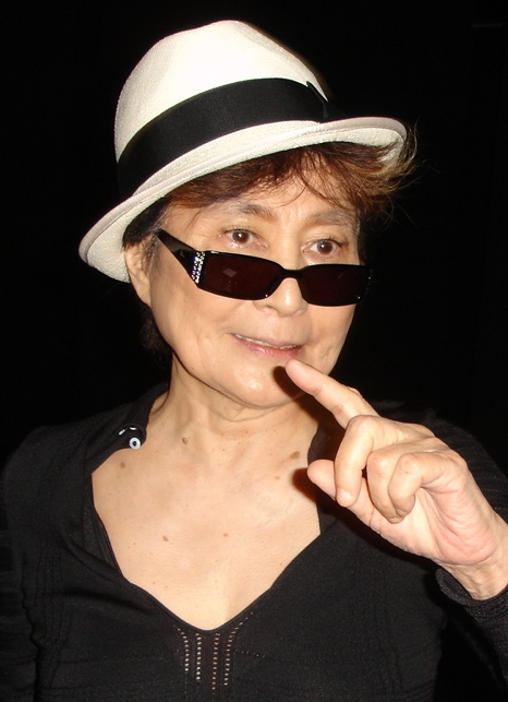 Yoko Ono mit einem weißen Strohhut und schwarzer Sonnenbrille. Sie zeigt mit dem Zeigefinger in Richtung der Kamera.