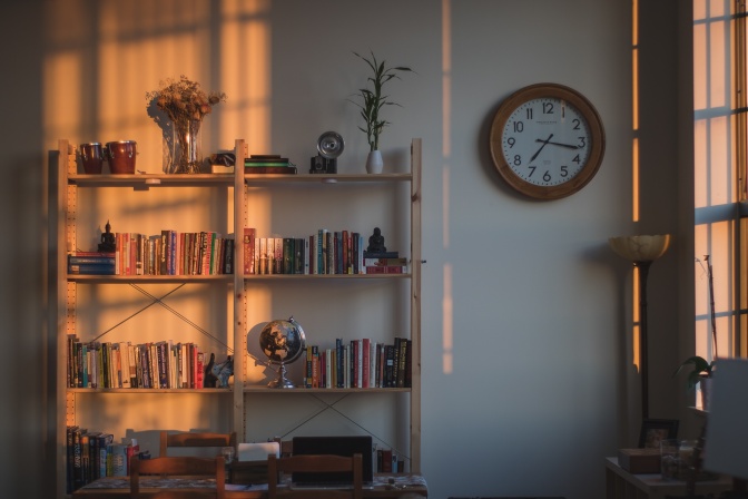 Ein Bücherregal in abnehmendem Abendlicht, das in den Raum fällt, Daneben hängt eine Wanduhr.