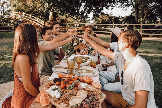 Eine Gruppe von Menschen sitzt an einem Tisch voller Essen und Getränke im Freien. Sie stoßen miteinander an.