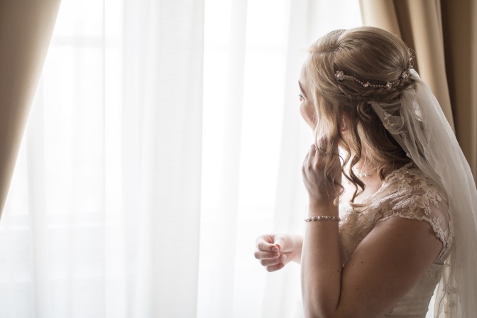 Eine Braut mit blonden, hochgesteckten Haaren und Schleier steht am Fenster und steckt sich einen Ohrring ins Ohr.