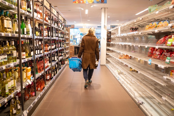Ein älterer Man im Ledermantel und mit langen, strähnigen weißen Haaren, geht mit einem Einkaufskorb aus Plastik durch den Supermarkt. Man sieht in von hinten zwischen Regalen mit Lebensmitteln auf beiden Seiten.