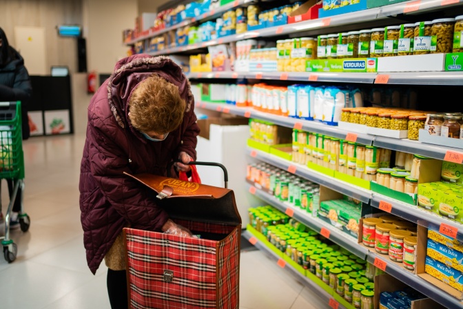 Eine alte Frau in einer Daunenjacke packt in einem Supermarkt Einkäufe in einen Einkaufstrolley.