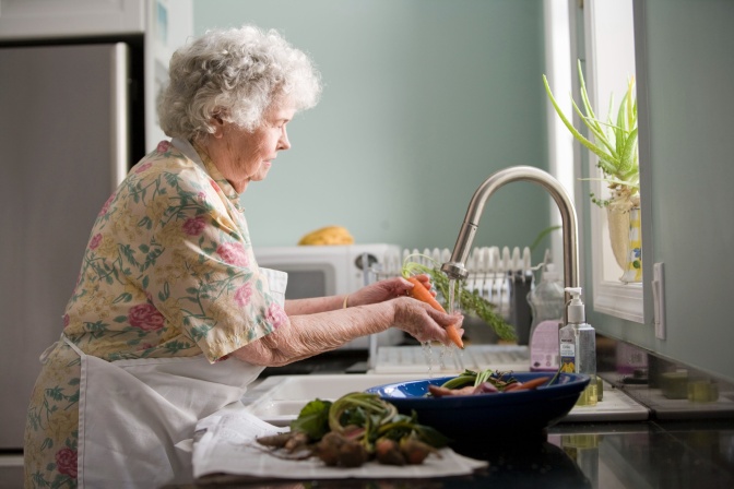 Eine alte Frau mit weißen Locken steht mit Schürze an einem Waschbecken in der Küche und wäscht Möhren.