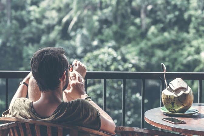 Ein Mann sitzt auf einem Balkon und hat die Füße aufs Geländer gestellt. Er schaut auf grüne Wälder. Neben ihm auf einem Tisch liegen eine Kokosnuss und ein Handy.