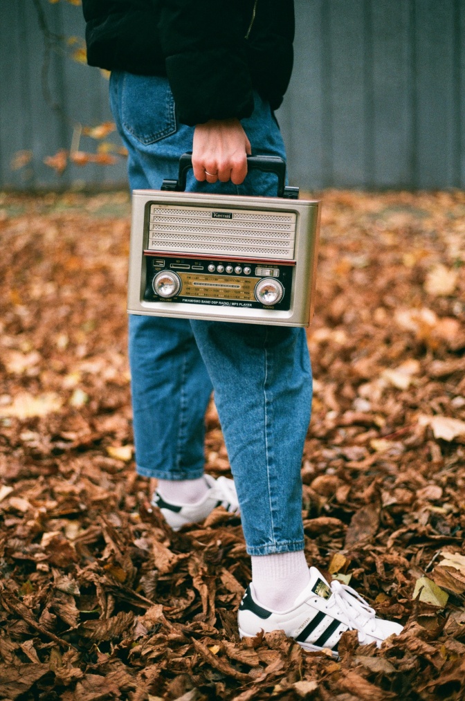 Eine Person in Senkern steht auf einem mit Blättern bedeckten Waldboden und hat ein altes Radio in der Hand.
