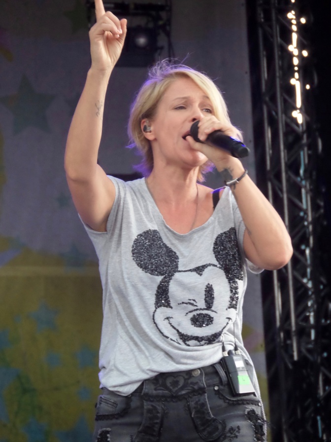 Michelle auf der Bühne. Sie singt in ein Mikro und streckt einen Arm hoch in die Luft. Sie trägt ein graues T-Shirt mit einer aus Paletten gestickten Micky Maus.
