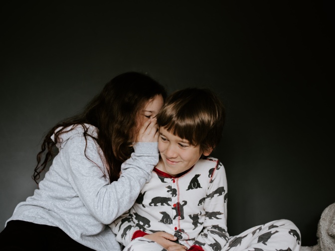 Ein Mädchen und ein Junge sitzen in Pyjamas zusammen. Sie flüstert ihn etwas ins Ohr.