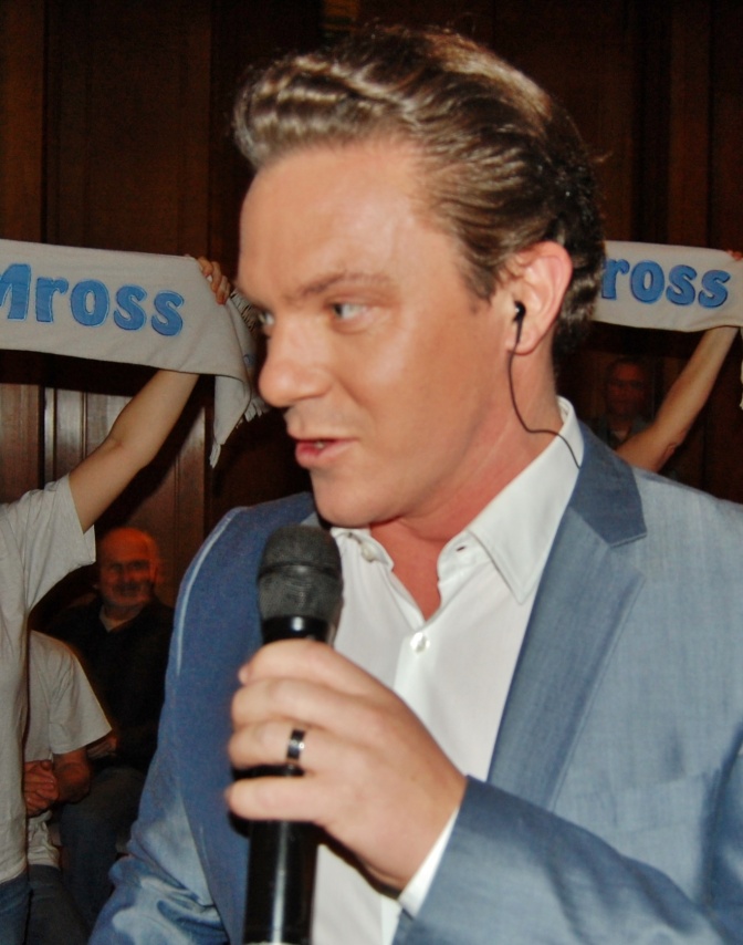 Stefan Mross in einem hellblauen Anzug mit gelockten blonden Haaren. Er singt in ein Mikrophon.