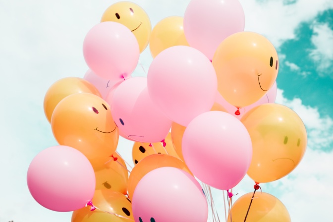Luftballons in orange und rosa. Auf den Ballons sind lächelnde Gesichter zu sehen.