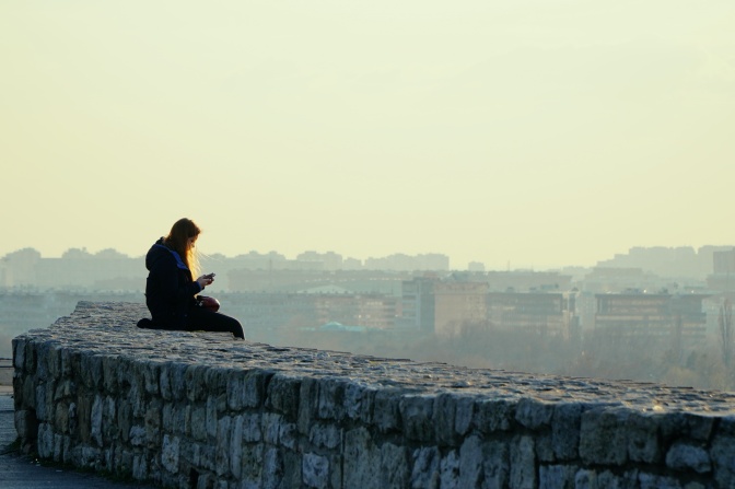 Eine Frau sitzt im Freien auf einer Mauer, im Hintergrund eine Stadt, die im Nebel liegt. Die Frau schaut auf ihr Handy.