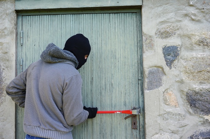 Ein Mann mit Strumpfmaske über dem Kopf bricht eine Holztür mit einem Stemmeisen auf.