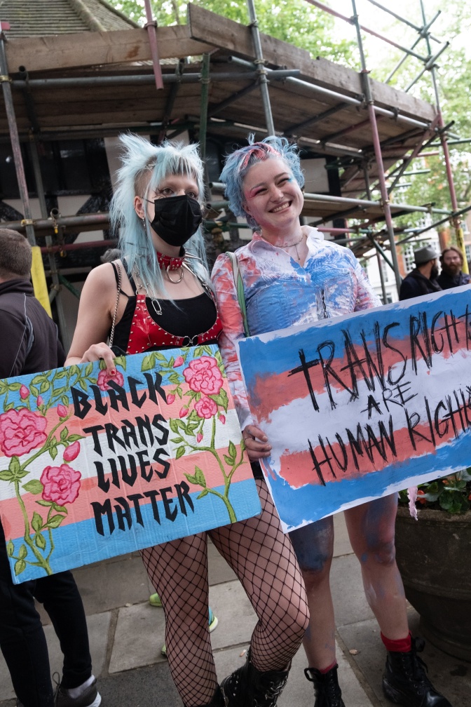2 Personen auf einer Demo. Sie haben ihre Haare hellblau und rosa gefärbt und tragen Protestschilder für trans Rechte.