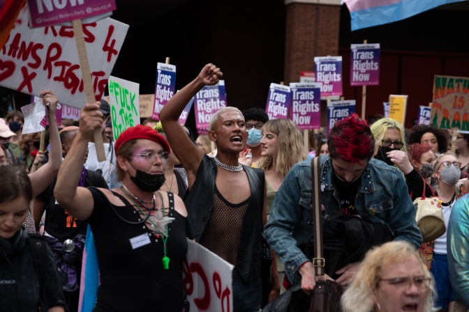 Eine Gruppe von trans Personen bei einer Demonstration. Einige von ihnen tragen Protestschilder, andere halten die Fäuste kämpferisch hochgestreckt.