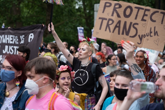 Eine Gruppe von trans Personen bei einer Demonstration. Einige von ihnen tragen Protestschilder, andere halten die Fäuste kämpferisch hochgestreckt. Manche von ihnen tragen Masken.