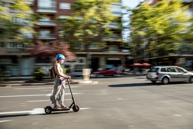 Ein Mädchen mit Rucksack fährt mit einem E-Scooter auf einer Straße. Der Hintergrund ist verschwommen, die Farben sind frühlingshaft.