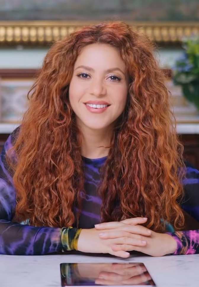 Shakira  mit langen, roten gelockten Haaren. Sie sitzt und hat die Hände vor dem Körper gefaltet. Sie schaut lächelnd direkt in die Kamera.