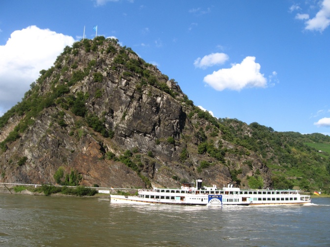 Ein hoher Felsen aus grauem Gestein am Ufer des Rheins. Teile des Felsens sind mit Bäumen bewachsen.