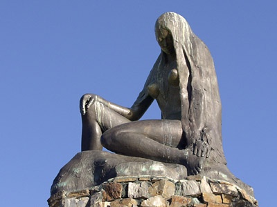 Eine sitzende Statue aus grauem Stein. Die nackte abgebildete Frau hat lange Haare und scheint in sich versunken zu sein.