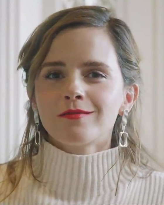 Emma Watson in einem hellen, gestrickten Rollkragenpullover und mit leuchtend rotem Lippenstift. Sie hat die dunkelblonden Haare zurückgebunden und trägt lange, hängende Ohrringe. Sie schaut direkt in die Kamera, ohne zu lächeln.