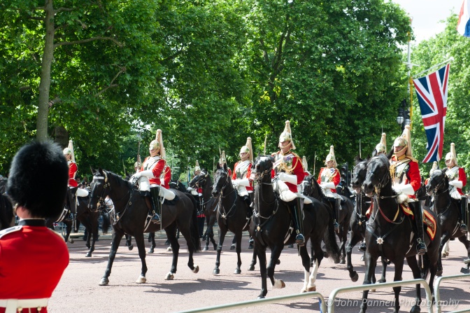 Britische Soldat*innen auf Pferden bei einer Parade. Sie tragen rote Uniformen und goldene Helme mit hohen Quasten.