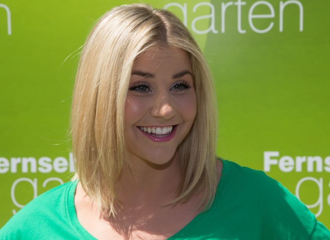 Beatrice Egli mit glatten, schulterlangen blonden Haaren. Sie steht lächelnd vor der Logowand des ZDF Fernsehgartens.