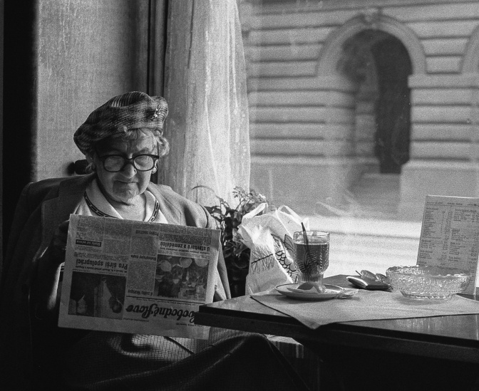 Schwarz-weiß-Foto einer alten Die, die in einem Café sitzt und Zeitung liest. Sie trägt Hut und Brille. Vor ihr auf dem Tisch steht ein Glas Tee.