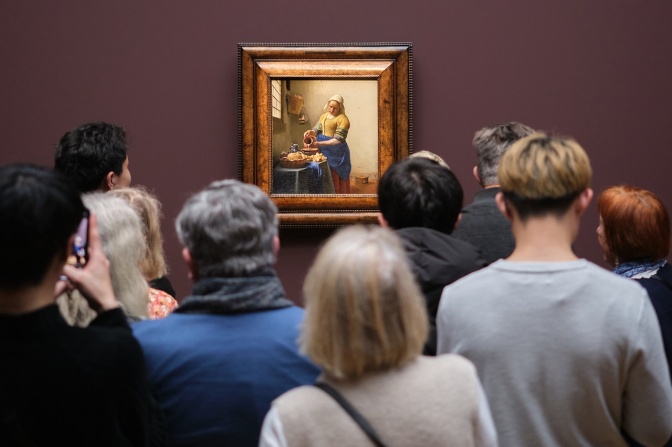 Eine große Gruppe von Menschen steht vor einem Gemälde in einem Museum. Das Gemälde zeigt eine Frau, die Wasser aus einem Krug gießt.