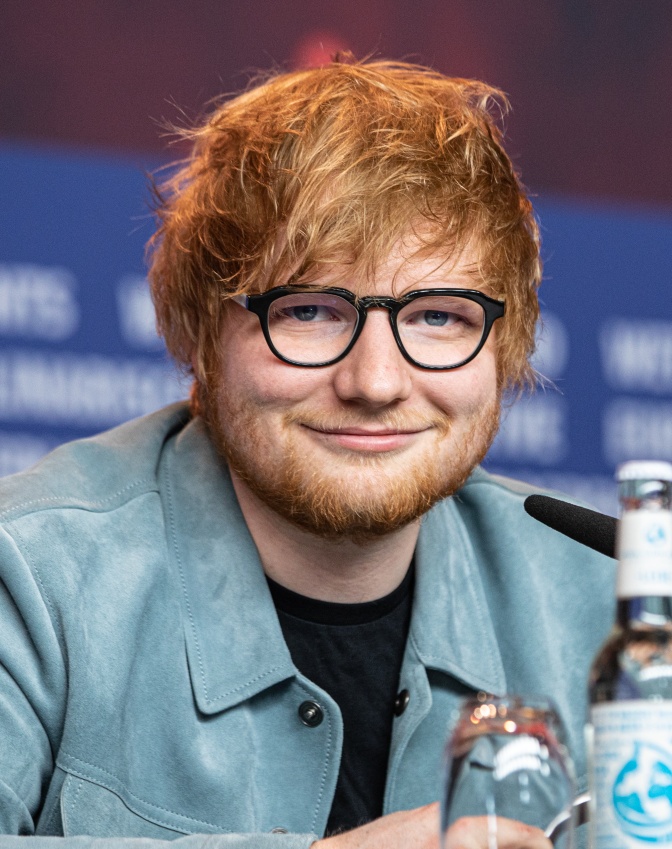 Ed Sheeran mit roten Haaren, Bart und Brille. Er trägt eine türkisfarbene Lederjacke und schaut direkt in die Kanera.