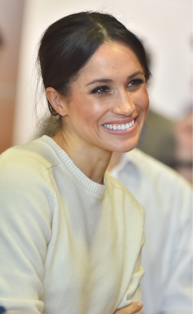 Herzogin Meghan mit langen, dunklen, zum Zopf gebundenen Haaren. Sie trägt einen weißen Pullover und lächelt.