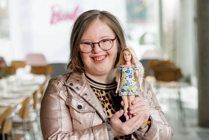 Eine Frau mit Down-Syndrom in Lederjacke und Leopardenshirt. Sie lächelt und hält eine Barbie in einem kurzen geblümten Kleid in der Hand.