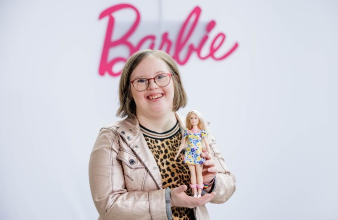 Eine Frau mit Down-Syndrom in Lederjacke und Leopardenshirt. Sie lächelt und hält eine Barbie in einem kurzen geblümten Kleid in der Hand. Im Hintergrund sieht man den pinkfarbenen Barbie-Schriftzug.
