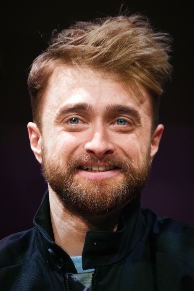 Daniel Radcliffe mit braunen, zur Seite frisierten Haaren und braunem Vollbart. Er lächelt und schaut seitlich an der Kamera vorbei.