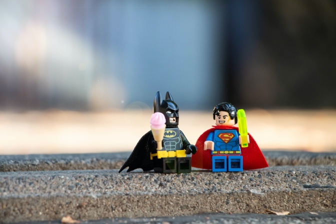 Eine Legofigur von Superman mit einem Laserschwert in der Hand und eine Legofigur von Batman mit einer Eistüte in der Hand.
