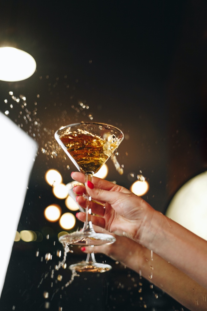 Zwei weibliche Hände mit rot lackierten Fingernägeln halten je einen Kelch mit Champagner in der Hand. Beide Gläser sind in Bewegung, es spritzt Champagner heraus.